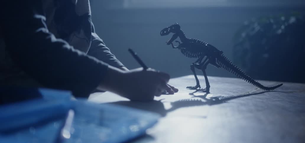 A boy making a sketch of a dinosaur skeleton toy in a dim room
