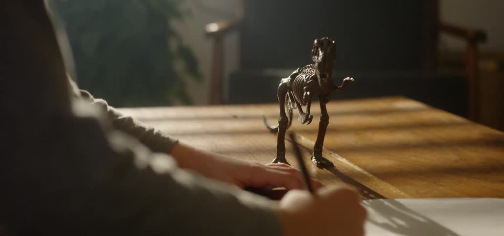 A child has a pencil and a piece of paper on a table beside a toy dinosaur