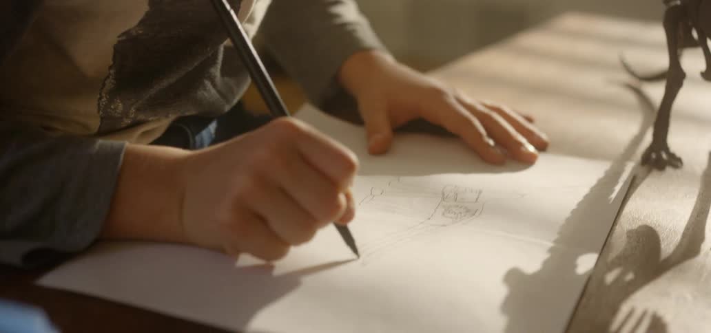 A young boy sitting at a table draws a picture of a dinosaur on a sheet of paper