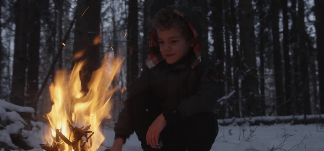 A young boy in a snowy forest warms himself next to a burning fire