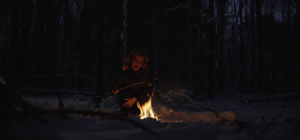 A young boy sits near a campfire and cooks food on the end of a long stick