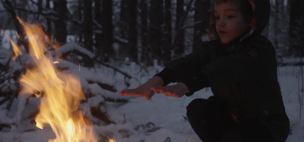 A young boy holds his hands above a campfire in the snow