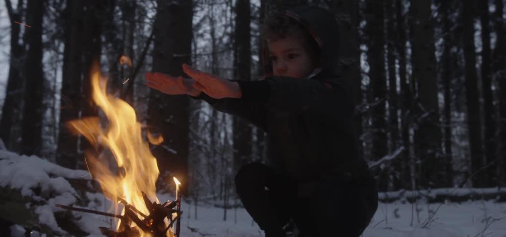 A young boy warms his hand over an open fire that is burning in a snow covered forest