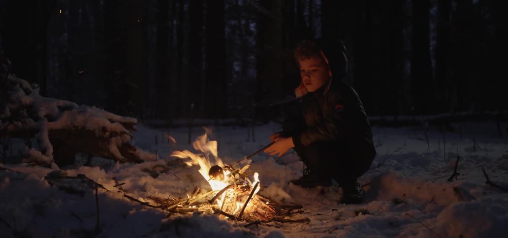 Child roasts apple over campfire while crouching on snow covered ground in woods