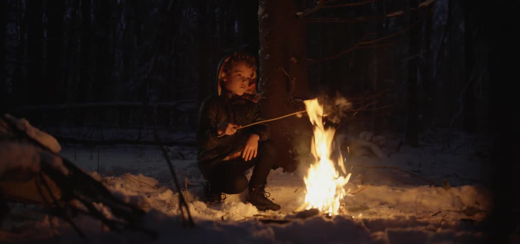 A boy roasts an apple on a stick over a campfire at night in a snowy forest