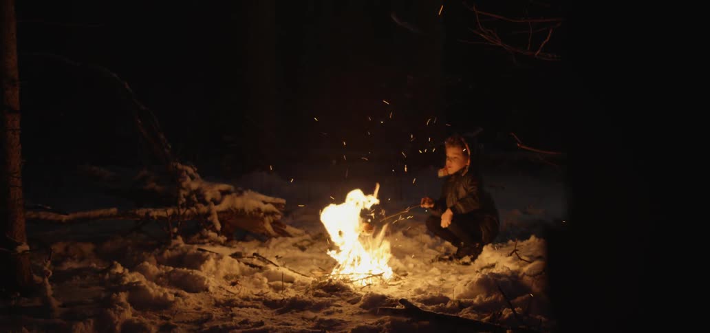 Boy crouches by campfire in snow roasting food