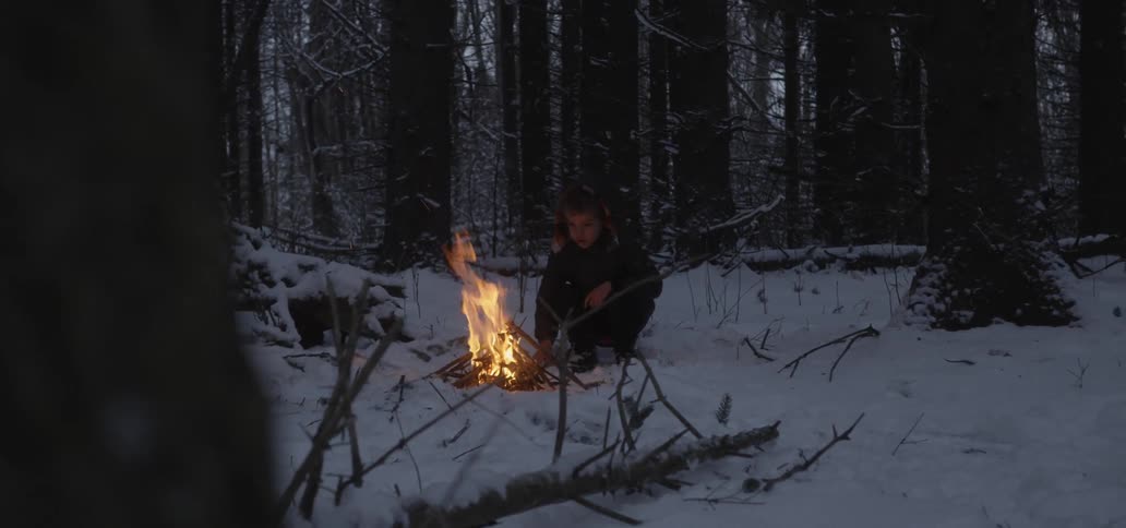 Boy crouches in snow to adjust campfire