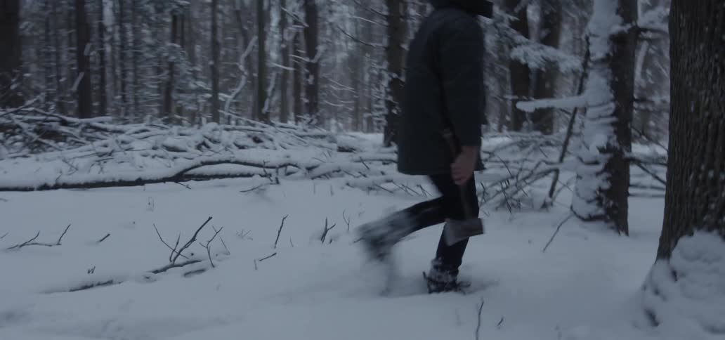 A boy walks through snow holding a small ax