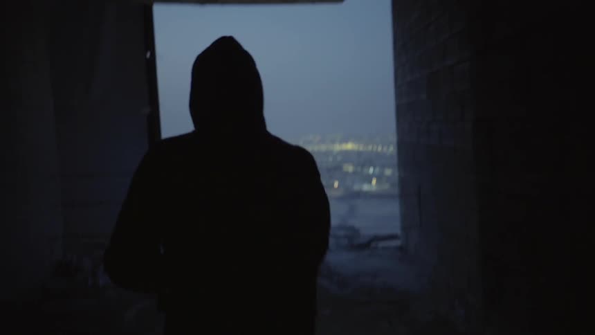 A man wearing a hoodie and holding a flashlight walks down a hall in an empty high-rise building until reaching an opening that looks out over a city at dusk