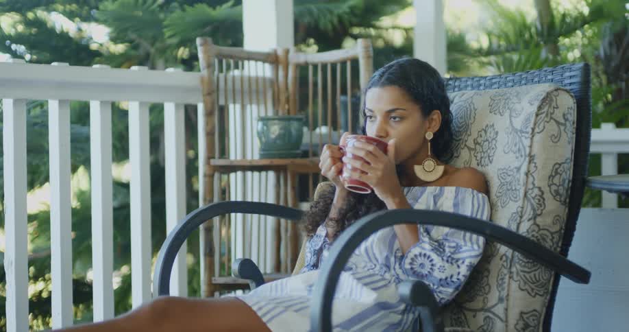 A woman sits down on a porch and drinks from a mug