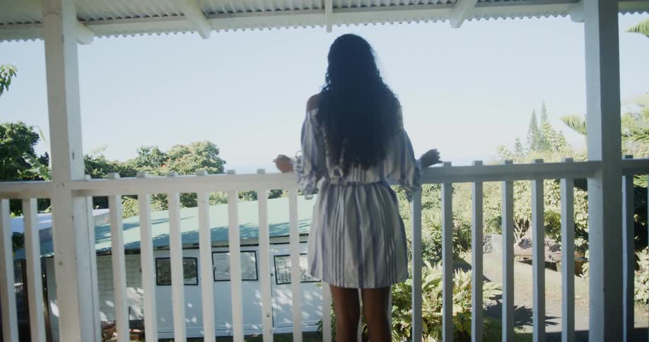 A young black woman in a summer dress walks outside on a balcony and looks out over the yard below