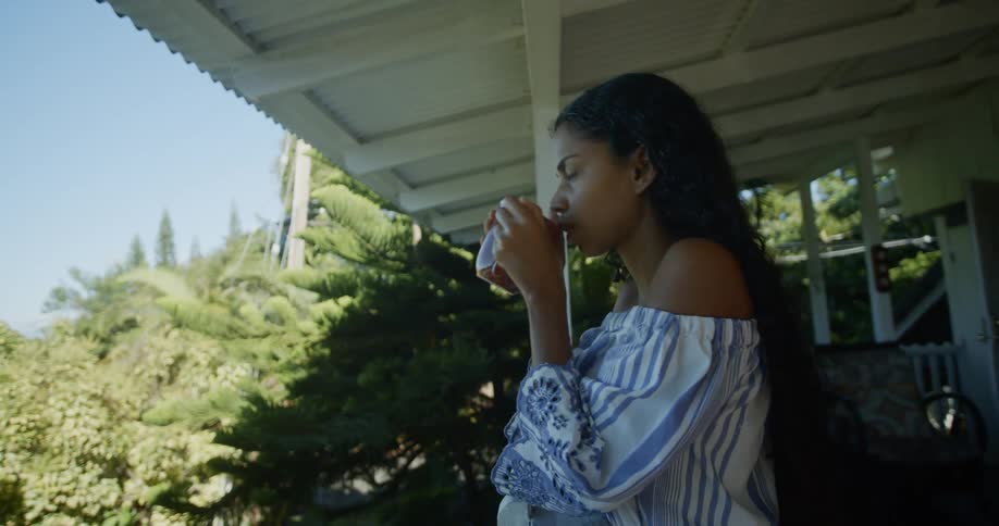 A woman drinking from a mug while staring out over a yard from a second floor balcony