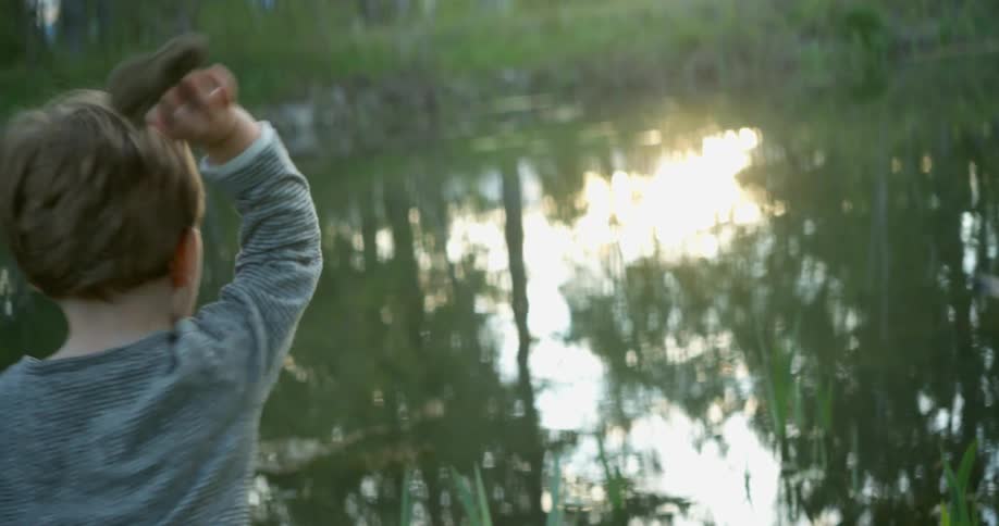 Toddler boy throws a stone into the lake at sunset
