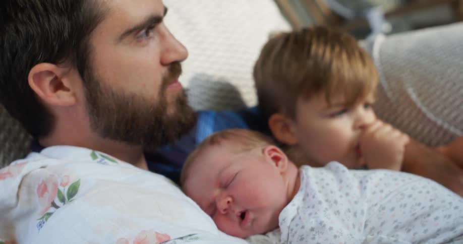A man sits on a couch with his son beside him and his baby sleeping on his chest