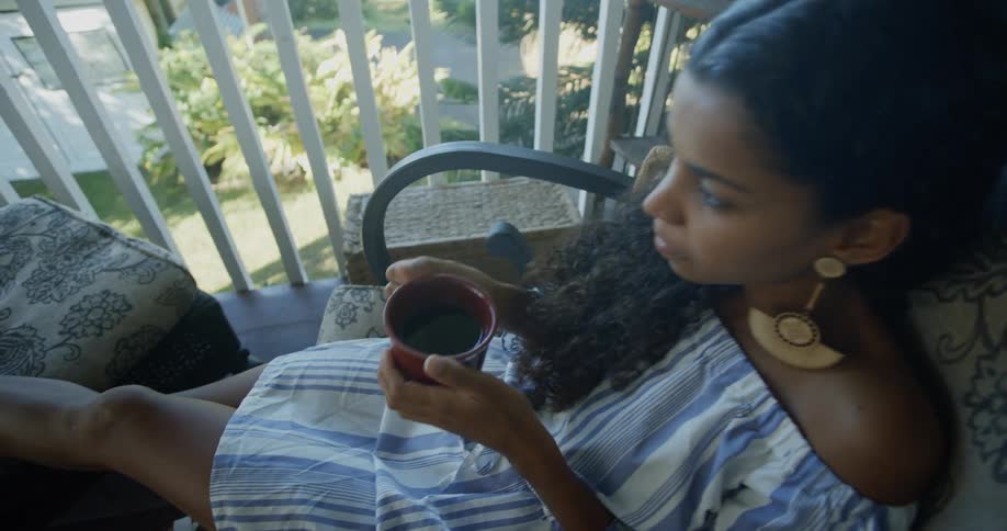 Young woman sits in a chair on a balcony and drinks something from the brown cup