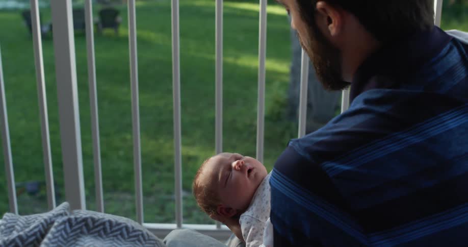 A dad sits outside holding his sleeping newborn in his hands