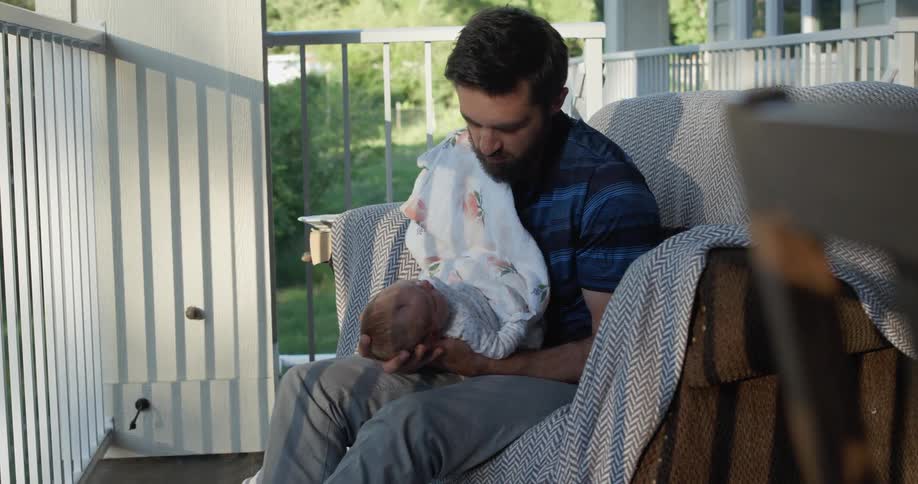 Father sitting on the couch holds and looks at the baby outdoors at sunset