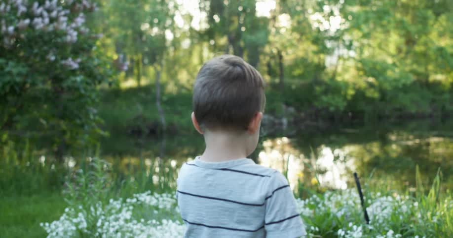 A toddler boy walks near flowers and a lake on a sunny day
