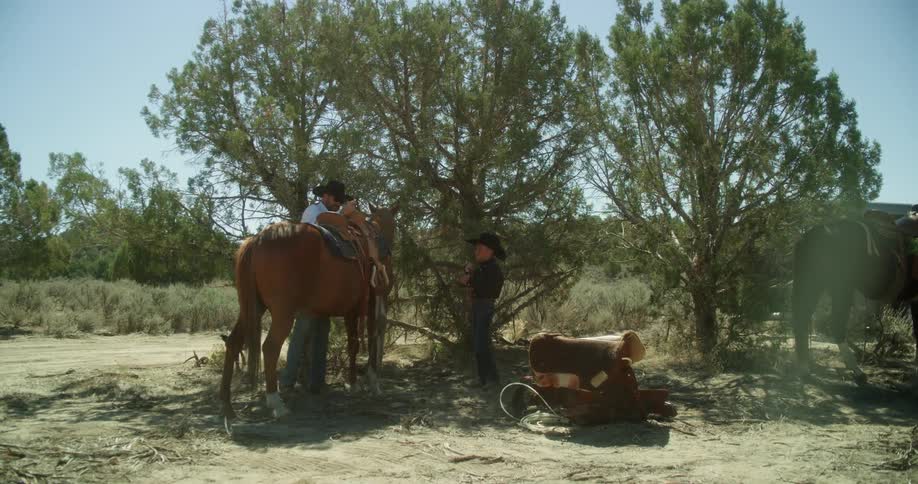 A man puts a saddle on a horse while a boy looks on