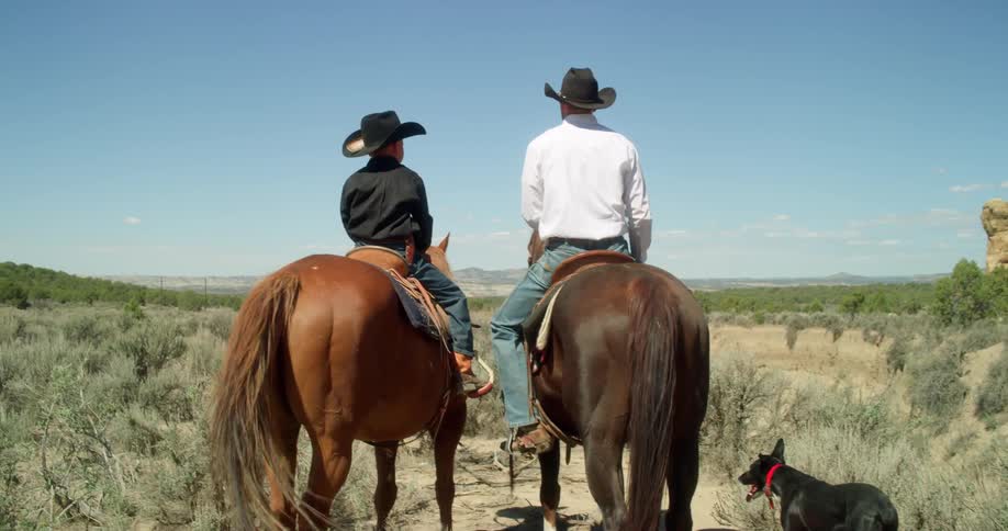 A man and a boy on horseback with their dog in an arid landscape