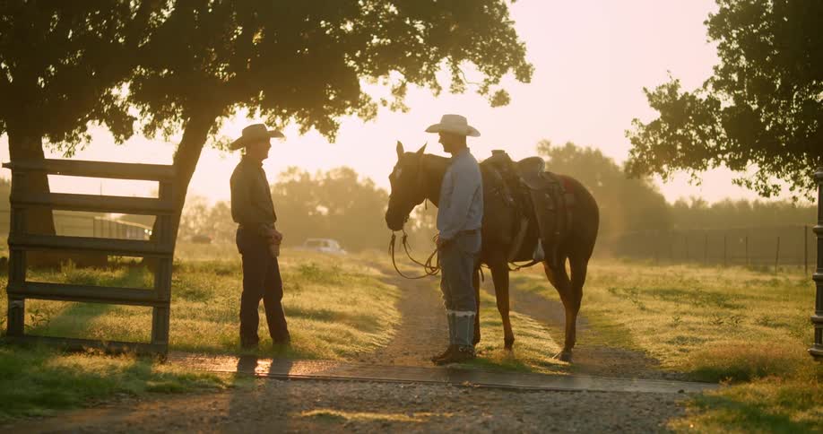 Two cowboys talk next a horse on a ranch at sunset