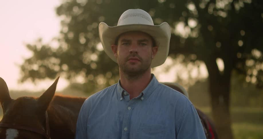 A cowboy stands next to a horse outside at sunset