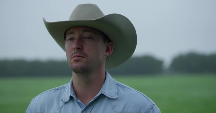 A man wearing a cowboy hat lifts his head while standing in a green field