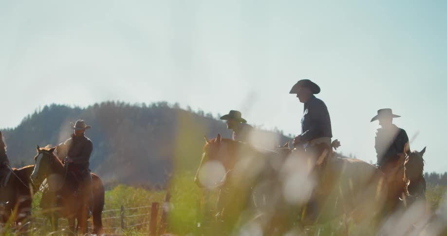 A group of men in cowboy hats sit atop horses that are standing idly in a field