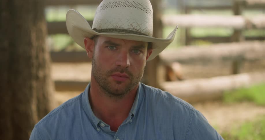 A cowboy stands in front of a wood fence on a ranch