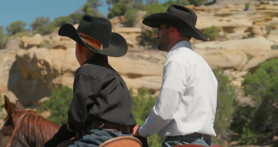 A dad talks to his son as they both sit on horseback looking at the rocky hills on a sunny day