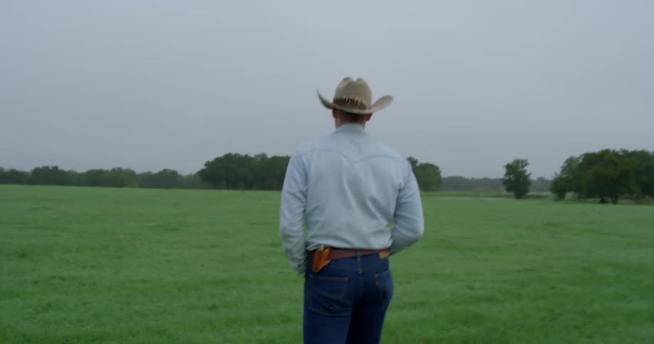 Man wearing a cowboy hat walks along the green meadow on a cloudy day