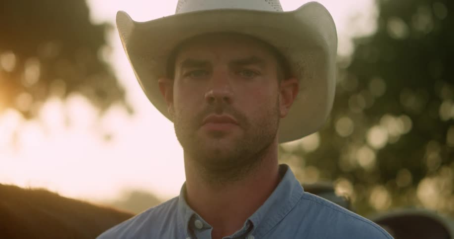 An adult male wearing a cowboy had stands next to a horse while looking into the camera