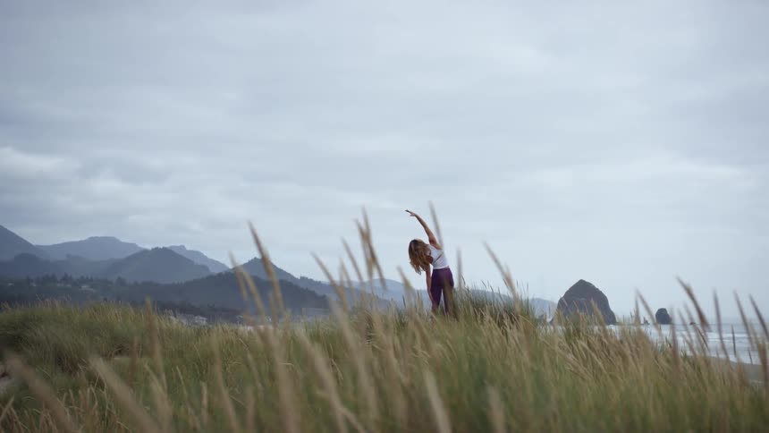 A woman stretches her arm high above her head as she stands outdoors in coastal area