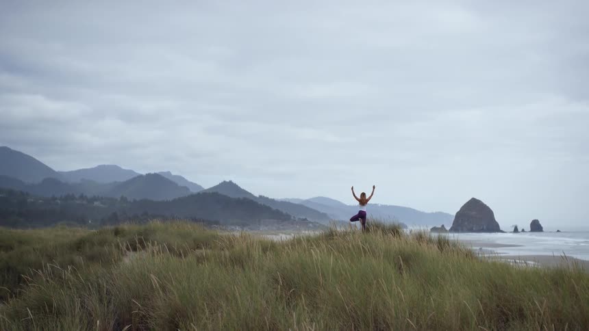 A woman is doing yoga on a grassy hill near the beach