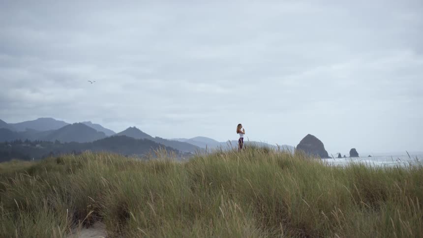 A woman stands on a hill in grasslands looking at the ocean with mountains in the background
