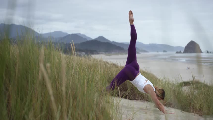 A woman at the beach practices yoga and holds a pose with her leg stretch high above her