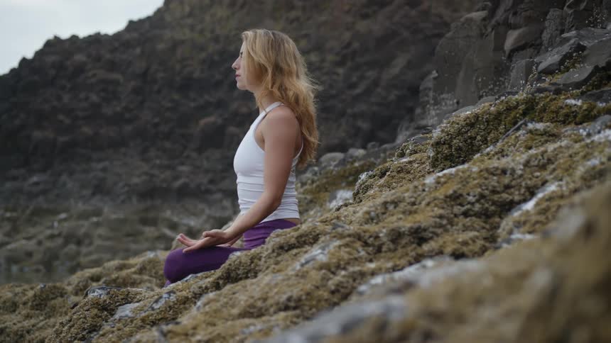 A woman in purple yoga pants meditates outside on a rocky surface