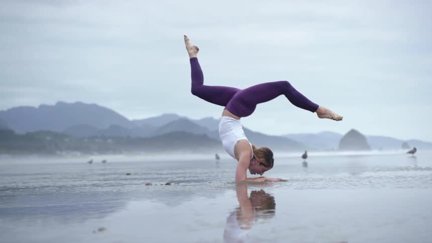 A woman is inverted as she performs a very challenging yoga pose on a wet sandy beach