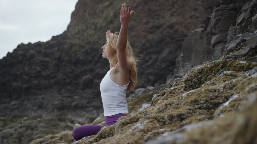 A woman sitting on the base of a rocky mountainside raises her hands above her head as she practices yoga