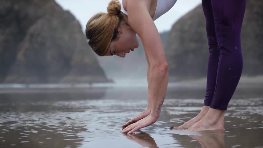 Woman performs yoga on wet beach near rocky hills