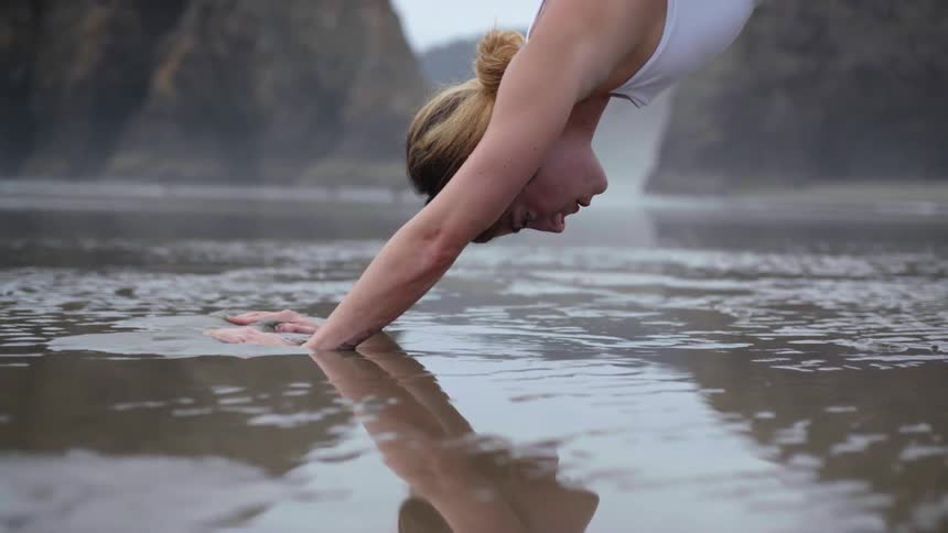 A woman stretching and holding a yoga pose in the wet sand is reflected on the water surface
