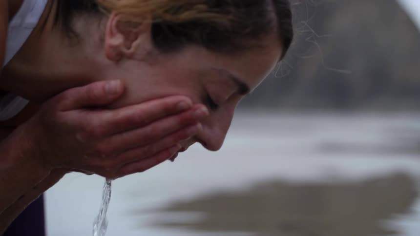 A woman leans downward to scoop up some water from a stream and wash her face with it