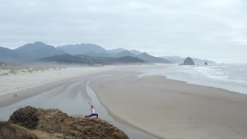 A woman practices yoga high atop a hill that overlooks the ocean and its beach