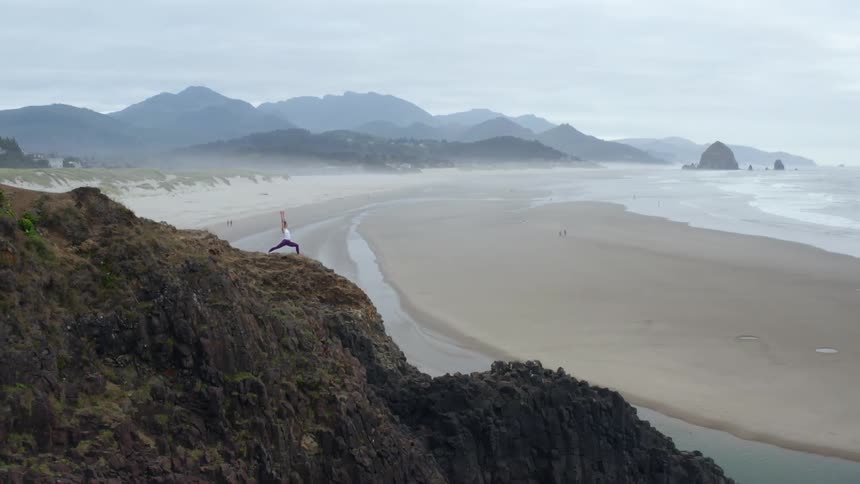 A woman stands high atop a cliff overlooking a coastal beach as she stretches her arms above her head in a yoga pose