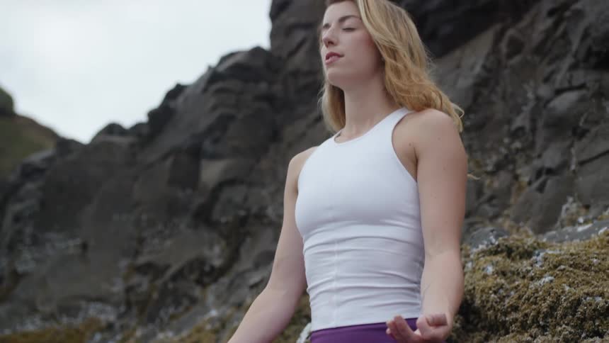 A woman in purple yoga pants meditates on mossy rocks