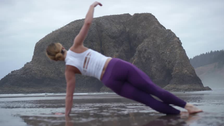 A woman lifts her arm above her head while holding a side plank pose in the wet sand