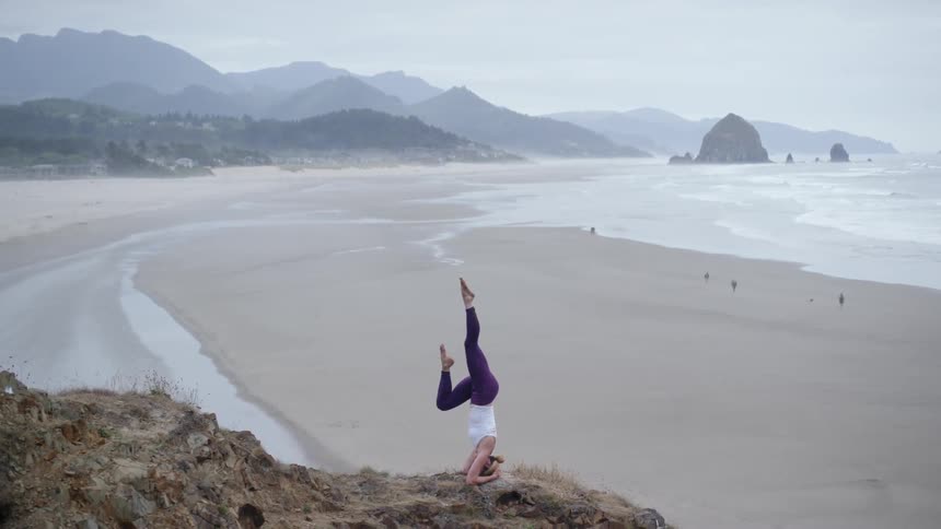 A white woman in a white shirt and purple yoga pants does yoga on high ground near a beach