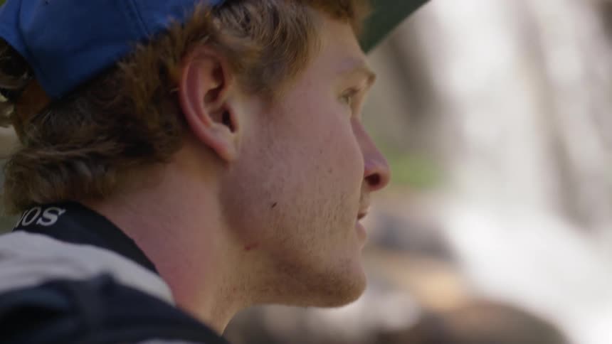 A young white man wearing a baseball cap sits outside near a waterfall