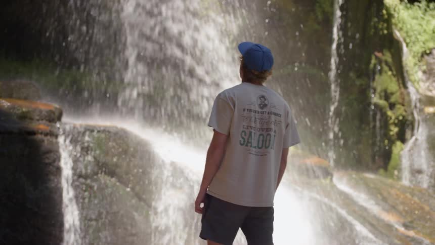 A man with a blue baseball hat standing in front of a waterfall