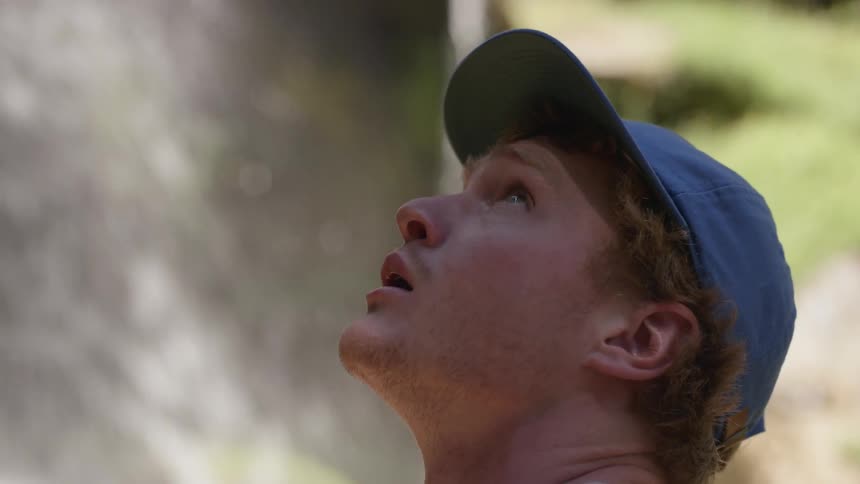 Blonde man wearing a blue cap looks at the waterfall in the forest